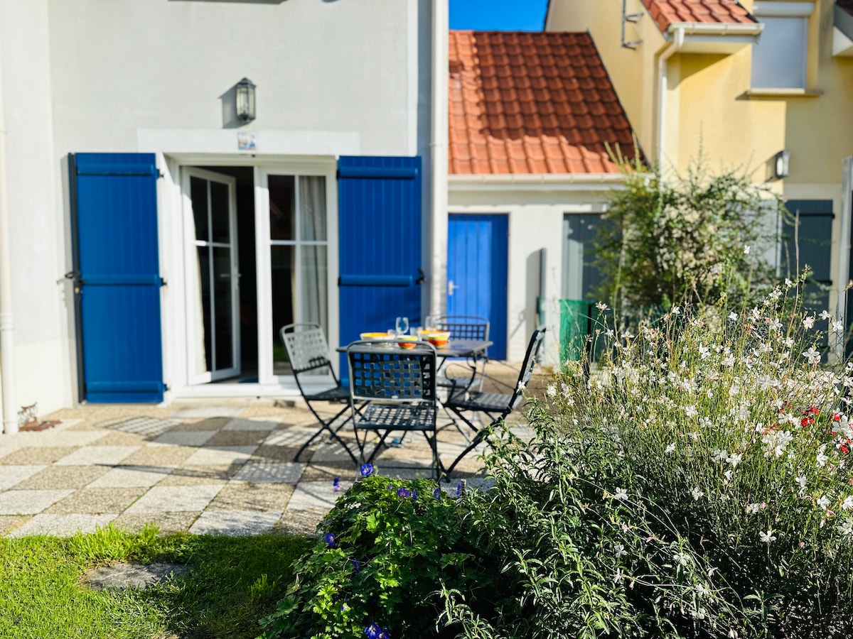 An outdoor terrace features a dining table and chairs, surrounded by blooming flowers and greenery. Blue shutters frame the entrance, complemented by a vibrant blue door in the background. The space is brightened by natural light, creating an inviting outdoor setting.