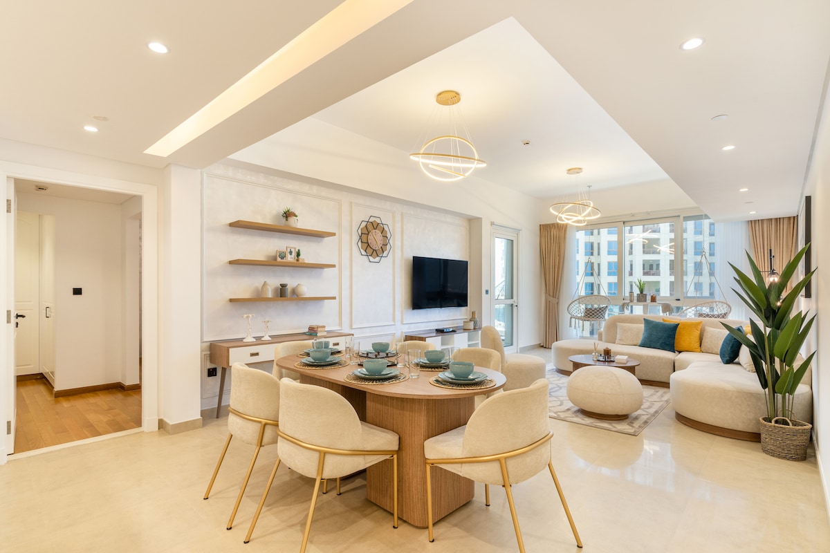 An inviting open-plan living space features a round dining table set for six, accented by stylish beige chairs. A plush seating area includes a large light-colored ottoman and colorful cushions. Natural light filters through expansive windows, complemented by soft neutral tones throughout the room.