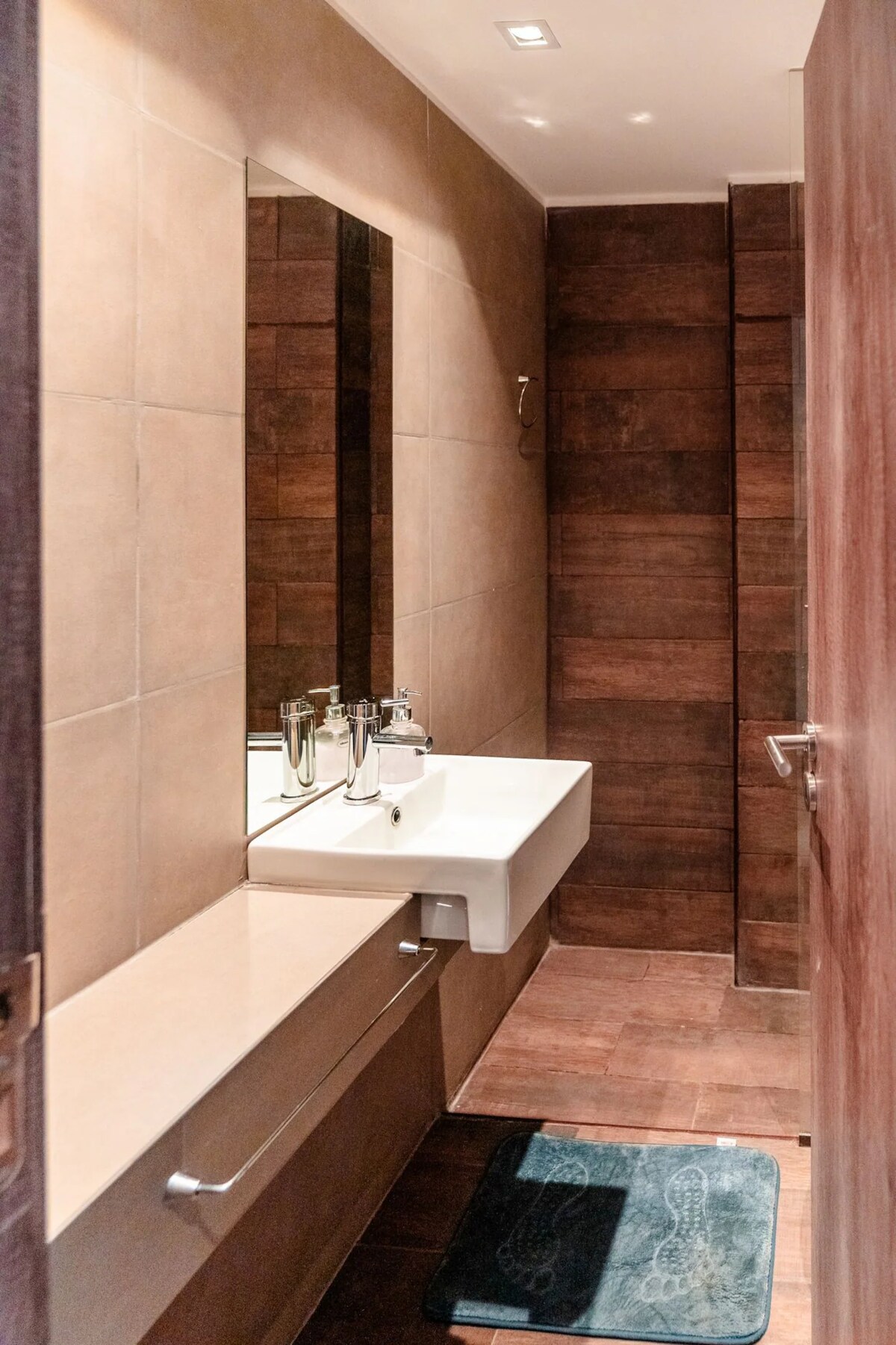 A compact bathroom features light-colored tiles and modern fixtures. A sleek, wall-mounted sink is positioned beneath a large mirror, while a beige countertop extends along the wall. Dark wooden accents create a warm contrast, complemented by a soft bath mat on the floor.