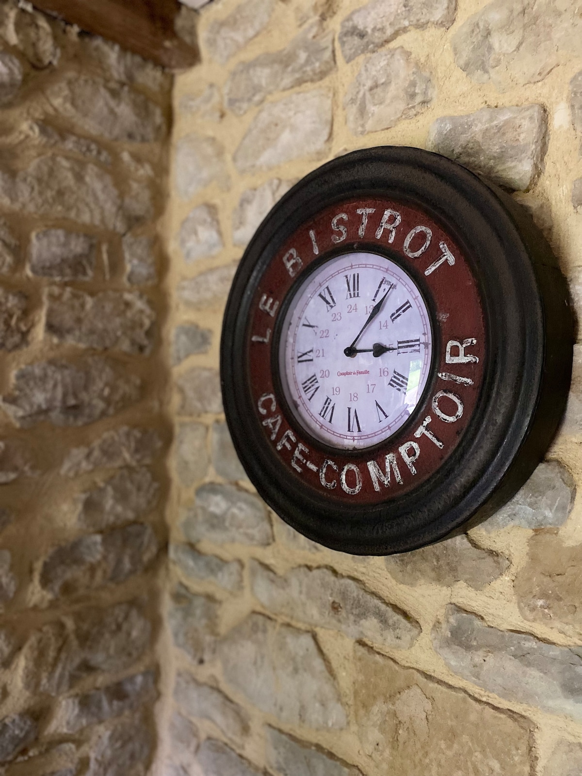 A round wall clock is mounted on a textured stone wall, featuring a dark red frame with the words 'Le Bistrot Café Comptoir.' The clock face displays classic numerals, providing a rustic charm to the space.