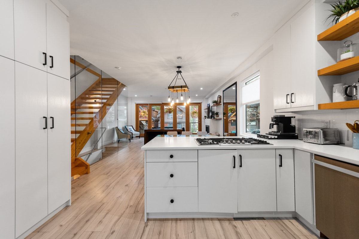The kitchen is designed with modern cabinetry and a spacious island with seating. A gas stove and coffee machine are visible. In the background, a dining area can be seen through large windows that offer natural light and views of the outdoor space. A staircase leads to the upper level.