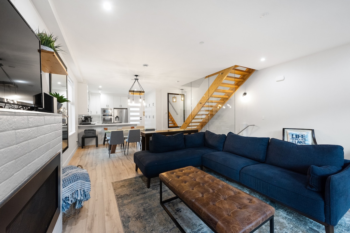 A spacious living area features a sectional sofa in a deep blue hue, paired with a tufted coffee table. A light wood dining table is visible in the background, along with a modern kitchen equipped with sleek appliances. Natural light enhances the open layout.