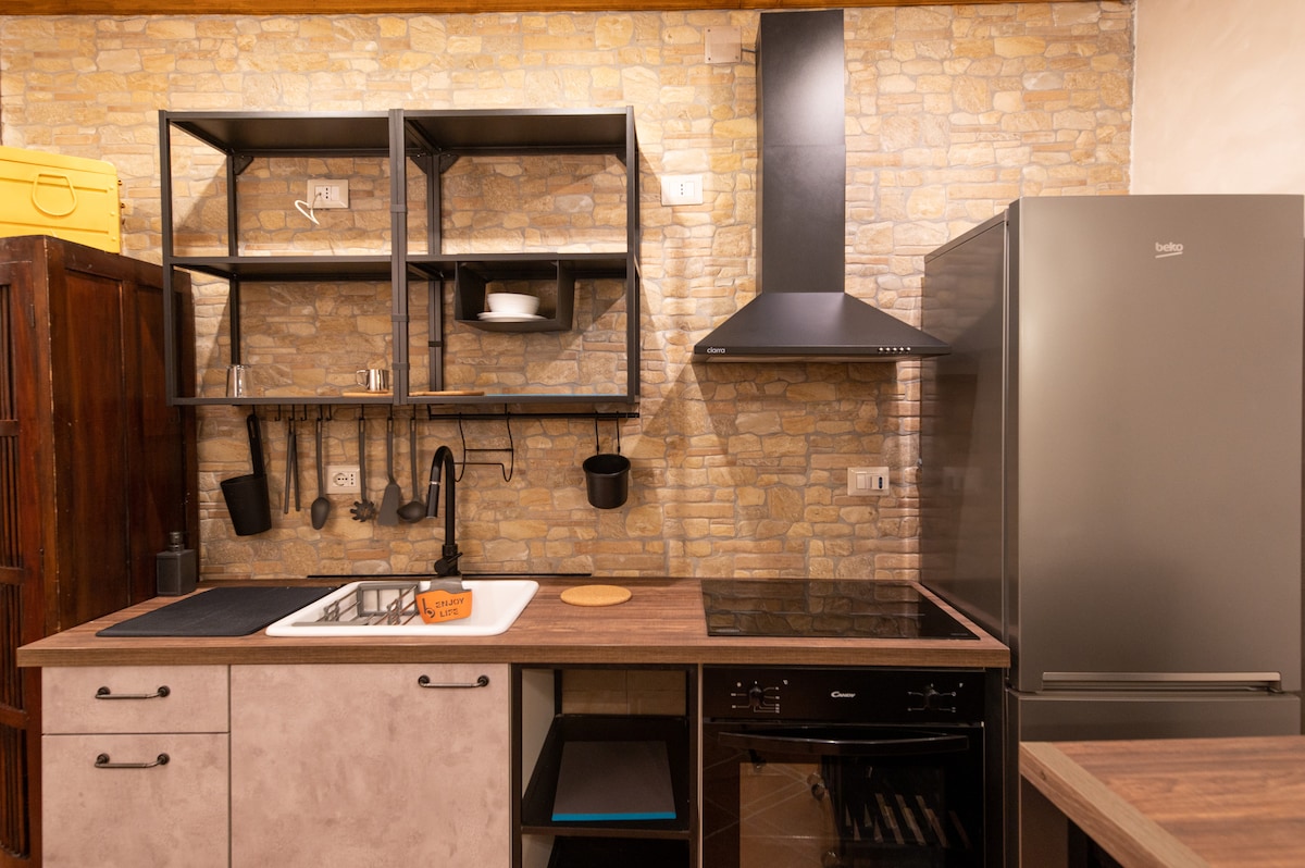 An industrial-style kitchen features open shelving, a modern stove, and a sleek black refrigerator. The countertop is complemented by a single-basin sink and a cutting board. Exposed stone walls add to the rustic charm, creating a functional cooking space.