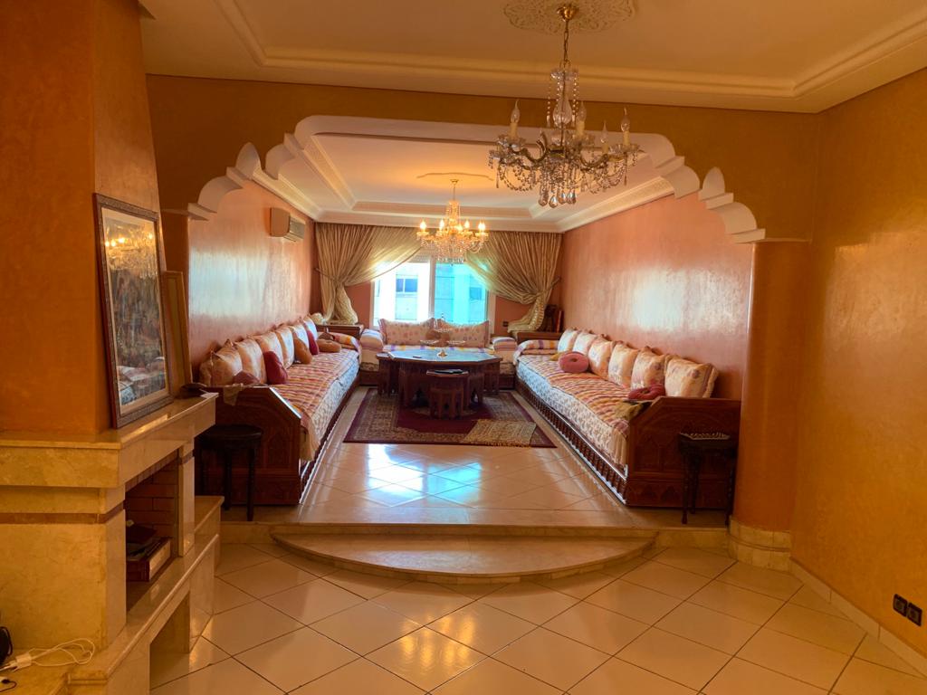A spacious living area features a large, U-shaped seating arrangement adorned with colorful cushions. A central coffee table invites gatherings, while elegant chandeliers hang from the ceiling. Natural light streams in through a large window, illuminating the warm tones of the walls.
