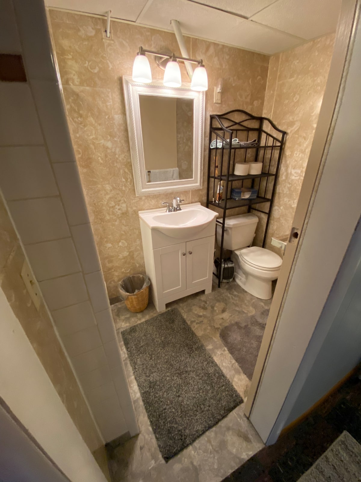 The bathroom features a white vanity with a sink, accompanied by a large mirror above it. A shelving unit holds neatly arranged items. The flooring is a muted grey, complemented by a dark mat near the sink. Natural light is provided by overhead fixtures.