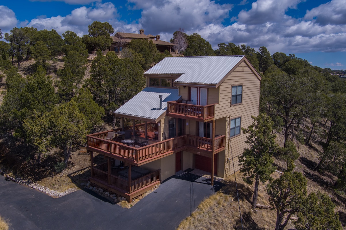 The exterior of the cabin is shown from an elevated angle, displaying two levels of spacious decks surrounded by trees. The siding is a light tan color with a metal roof. A paved driveway leads to the entrance, ensuring easy access.