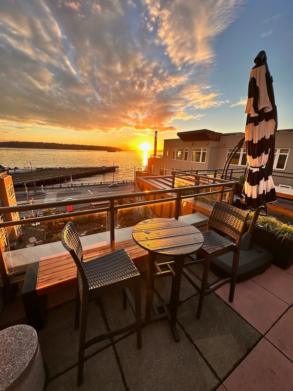 A rooftop area features a circular table accompanied by two dark chairs, set against a stunning sunset over the water. An umbrella stands nearby, adding to the inviting outdoor space that overlooks the scenic waterfront.