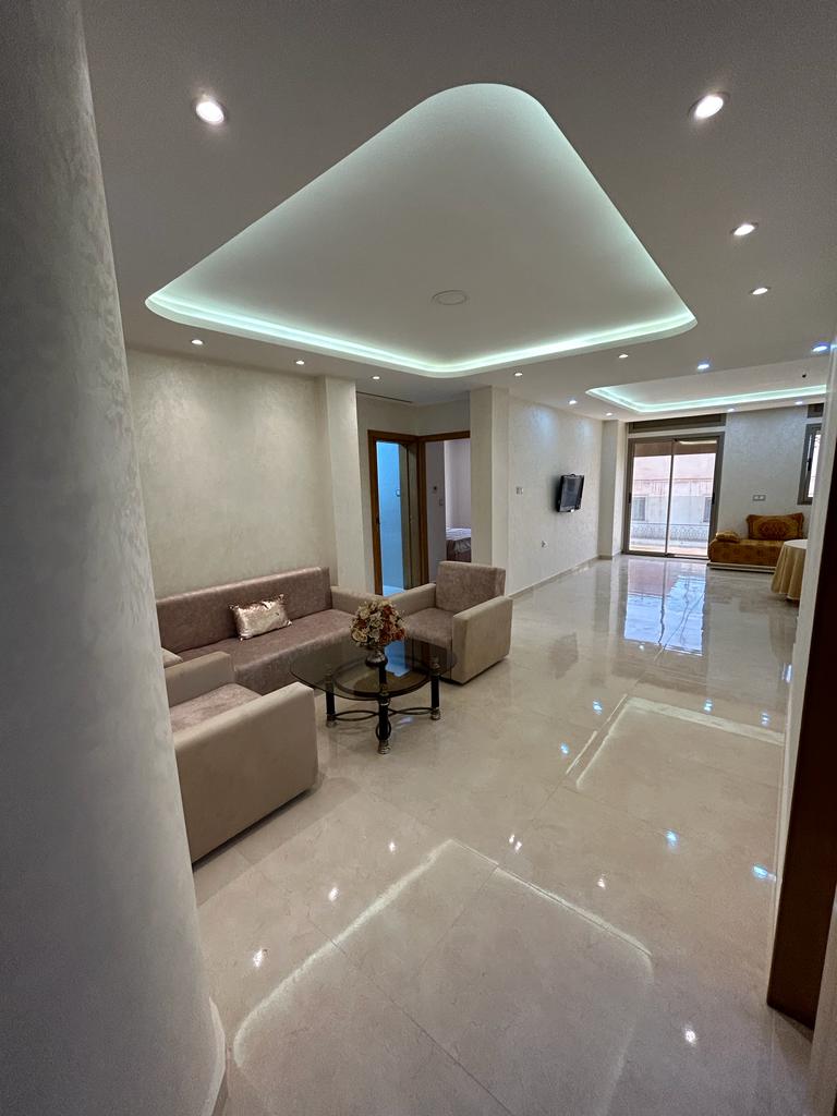 A modern living area features a light-colored sofa set arranged around a dark coffee table. Soft overhead lighting highlights the sleek, glossy floor. Large windows allow natural light to fill the space, creating an open and airy atmosphere.