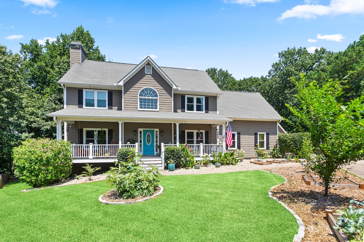 A spacious and welcoming home features a front porch with white railings, complemented by a vibrant blue front door. Lush green grass and landscaped gardens enhance the curb appeal, while a flag hangs proudly beside the entrance, reflecting a sense of community.