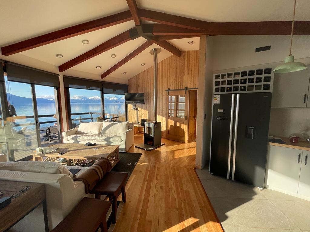 An inviting living area features large windows framing stunning views of the lake and mountains. The wooden ceiling beams add character, while a cozy seating arrangement includes a light-colored sofa and wooden furniture. A modern kitchen area is visible, showcasing sleek appliances.