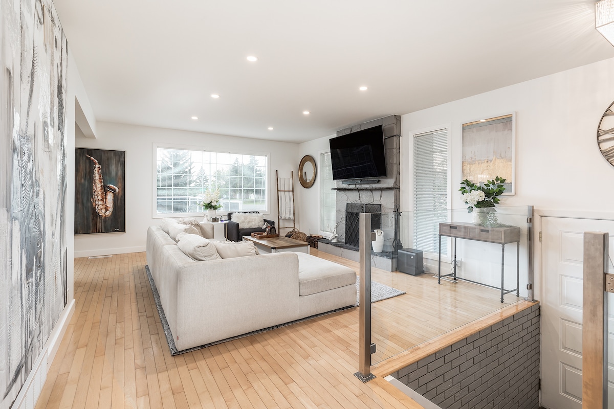 A spacious living area is highlighted by a large sectional sofa, complemented by a light wood floor. A wide window allows natural light to fill the room, while a mounted flat-screen TV is visible above a sleek console table. Minimalist decor elements create a contemporary feel.