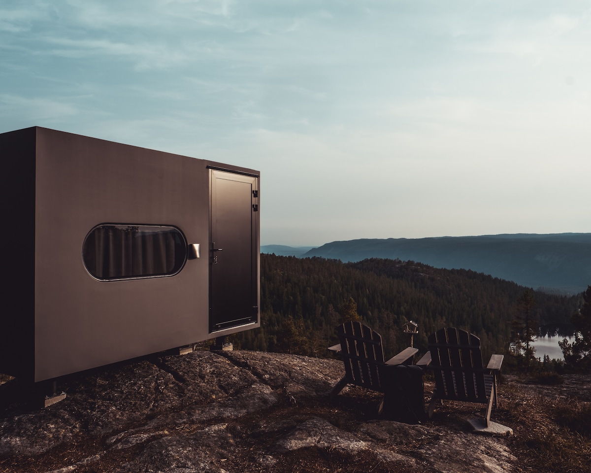 A modern structure is positioned on a rocky outcrop, offering expansive views of the surrounding forested hills. Two wooden chairs are situated nearby, inviting relaxation. The exterior features a large, rounded window and a sleek door, blending contemporary design with the natural landscape.