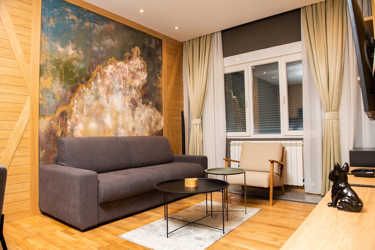 A modern living area features a dark gray sofa positioned alongside a light-colored chair. A round black table sits in the center on a textured rug, while a large abstract painting adorns the wall, creating a focal point. Natural light streams in through the windows, covered by sheer curtains.