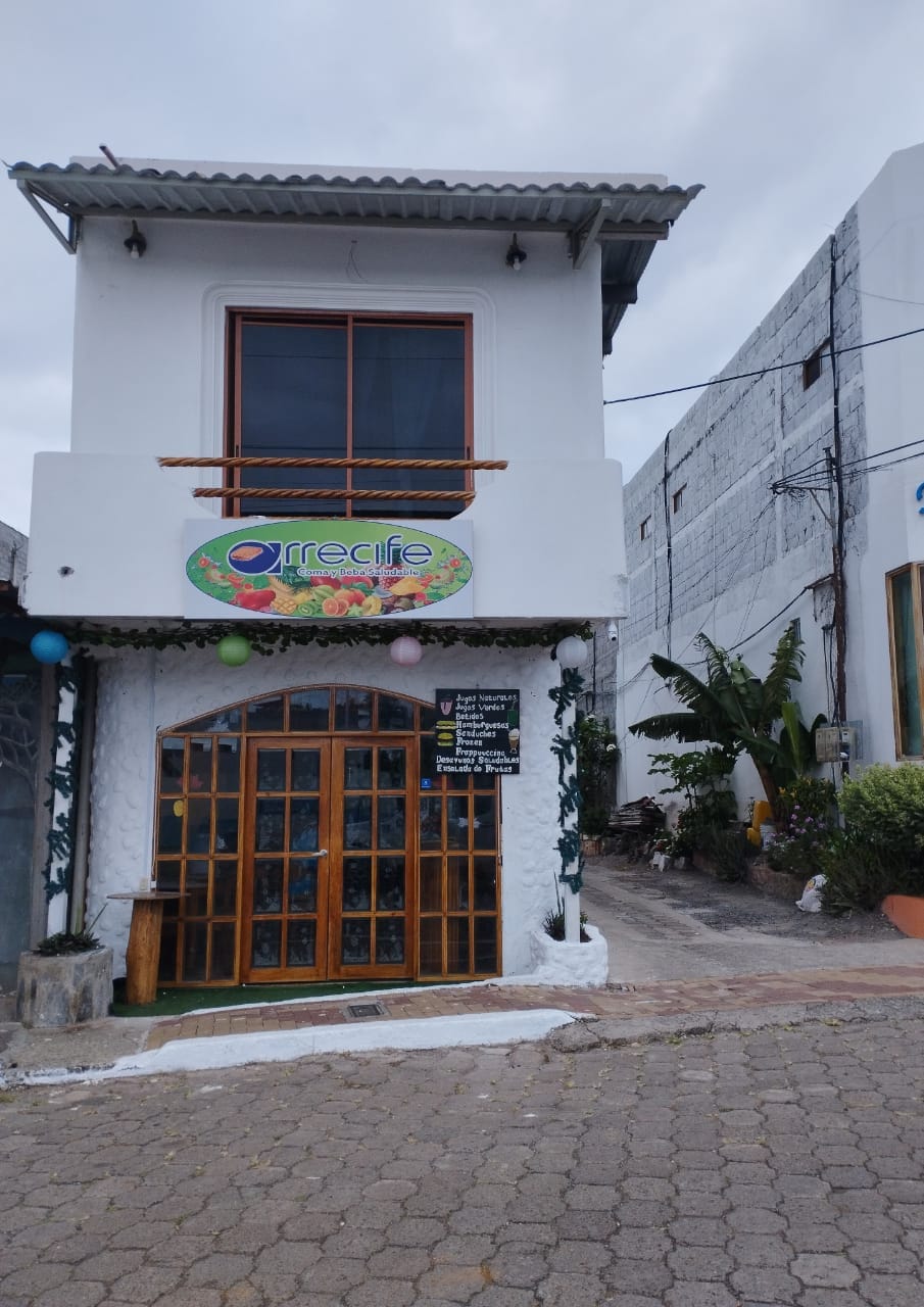 A two-story building is featured, characterized by a large window with wooden framing. The ground floor has double doors made of wood and glass, adorned with decorative elements. Tropical plants are visible along the pathway, contributing to a welcoming entrance.