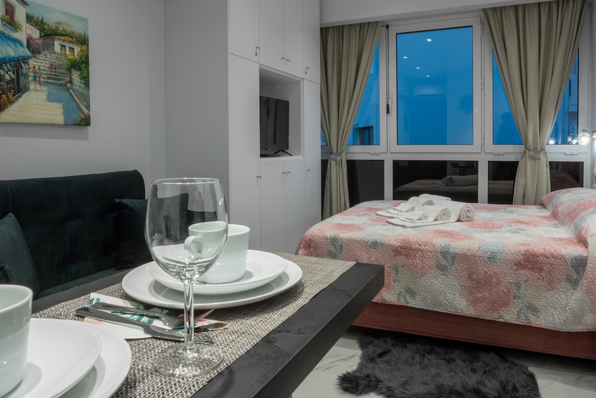 A bright interior is seen, featuring a cozy sleeping area with a neatly made bed adorned with a pastel quilt. A dining table is set with white dishware and glassware, while large windows overlook the city, complemented by light curtains that softly frame the view.
