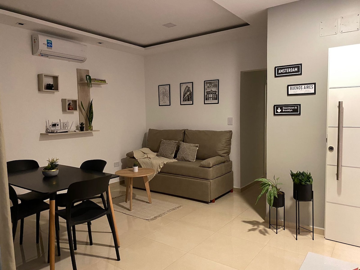 A modern living area features a light-colored tile floor and soft neutral walls. A cozy sofa is positioned beside a small coffee table, with a dining table set for four nearby. Green plants add a touch of nature to the space, complemented by framed wall art.