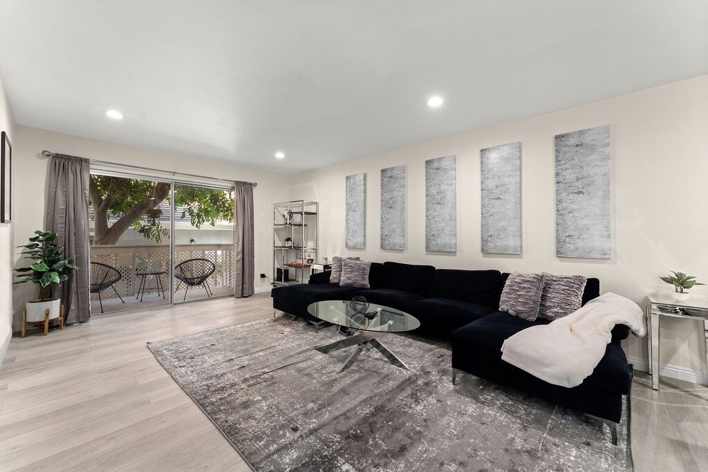A spacious living area features a large black sectional sofa and a geometric glass coffee table. Abstract art pieces are arranged on the light-colored walls. Natural light streams in through sliding glass doors that lead to a balcony, enhancing the calming atmosphere.