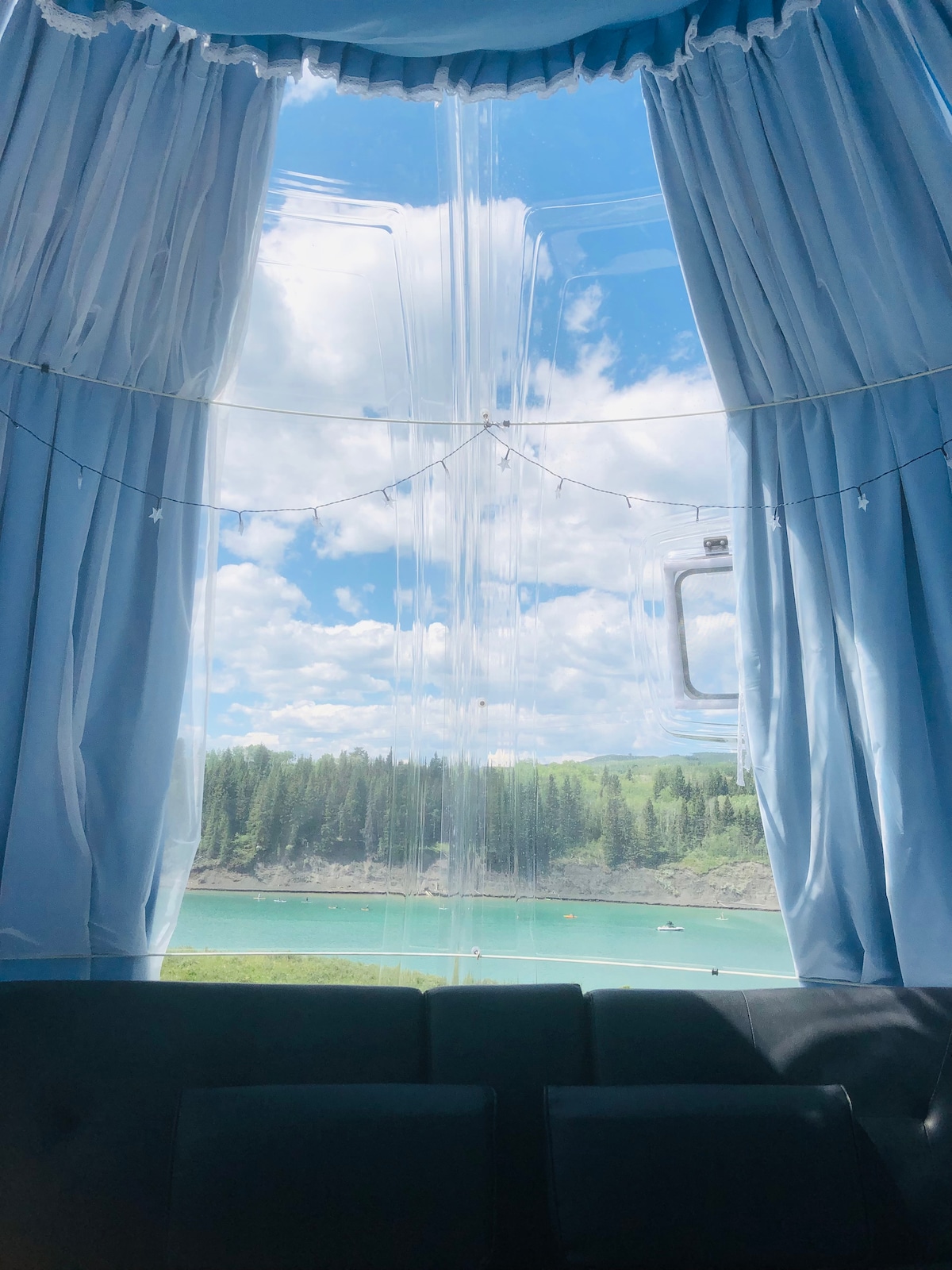 A large window framed by soft light blue curtains provides a view of the serene waters and lush greenery. Natural light illuminates the cozy interior, creating a refreshing and inviting atmosphere.