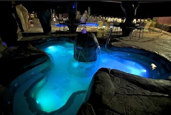 A jacuzzi is set within a stone enclosure, showcasing sparkling blue water illuminated by soft lighting. Surrounding seating is provided by black metal chairs, while a serene outdoor area is visible in the background, adding a tranquil atmosphere for relaxation.