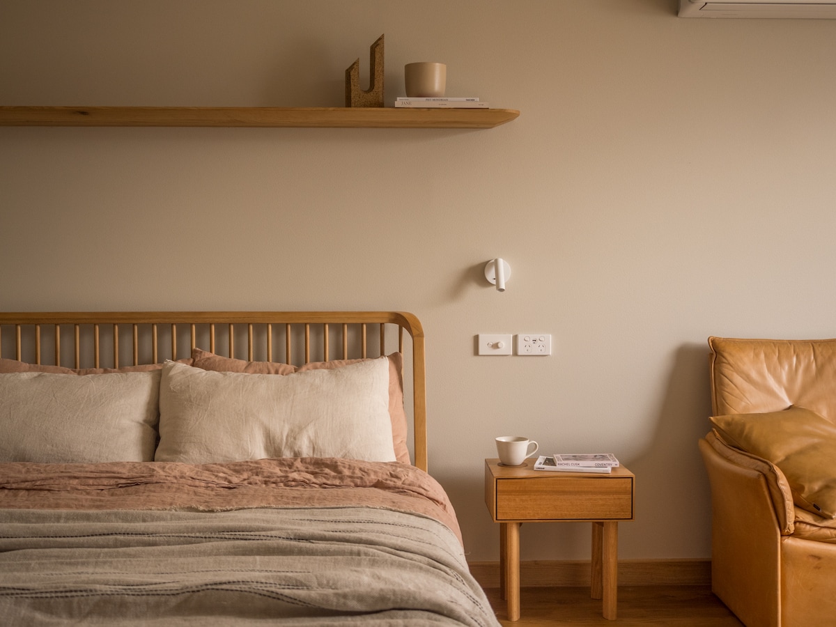 A cozy sleeping area features a queen-sized timber bed adorned with linen bedding in soft tones. A wooden nightstand holds a cup and a magazine. A rustic leather chair adds warmth, and a minimalist shelf displays a decorative item, enhancing the serene atmosphere.