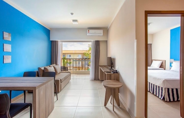 An inviting living area features a comfortable sofa and a stylish wooden table, complemented by a blue accent wall. Natural light flows through the large window, which offers views of the balcony. The adjoining bedroom area is partially visible, showcasing two beds.