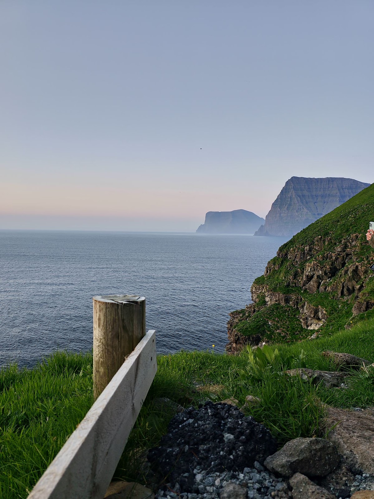 A coastal view captures steep cliffs meeting the calm sea, with distant mountains partially shrouded in mist. A wooden railing and a patch of grass add depth to the serene landscape, framed by the soft hues of the sky at dusk.