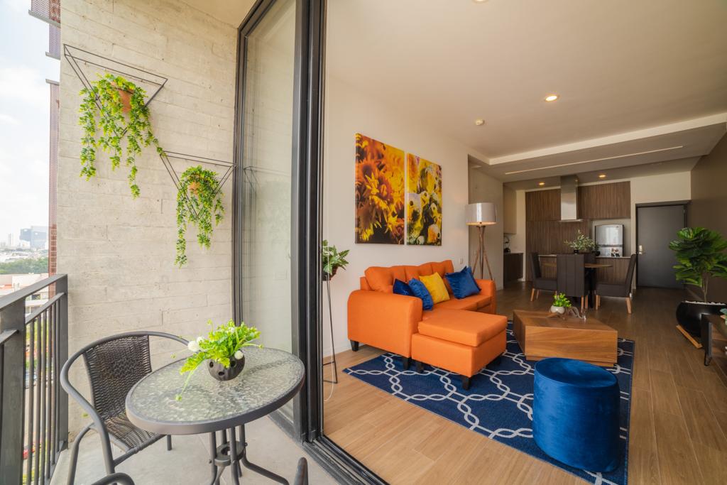 A spacious living area features an orange sofa accompanied by a round coffee table and a blue pouf. A balcony table with two chairs overlooks the surroundings. Bright floral artwork adorns the wall, and a kitchenette is visible in the background.