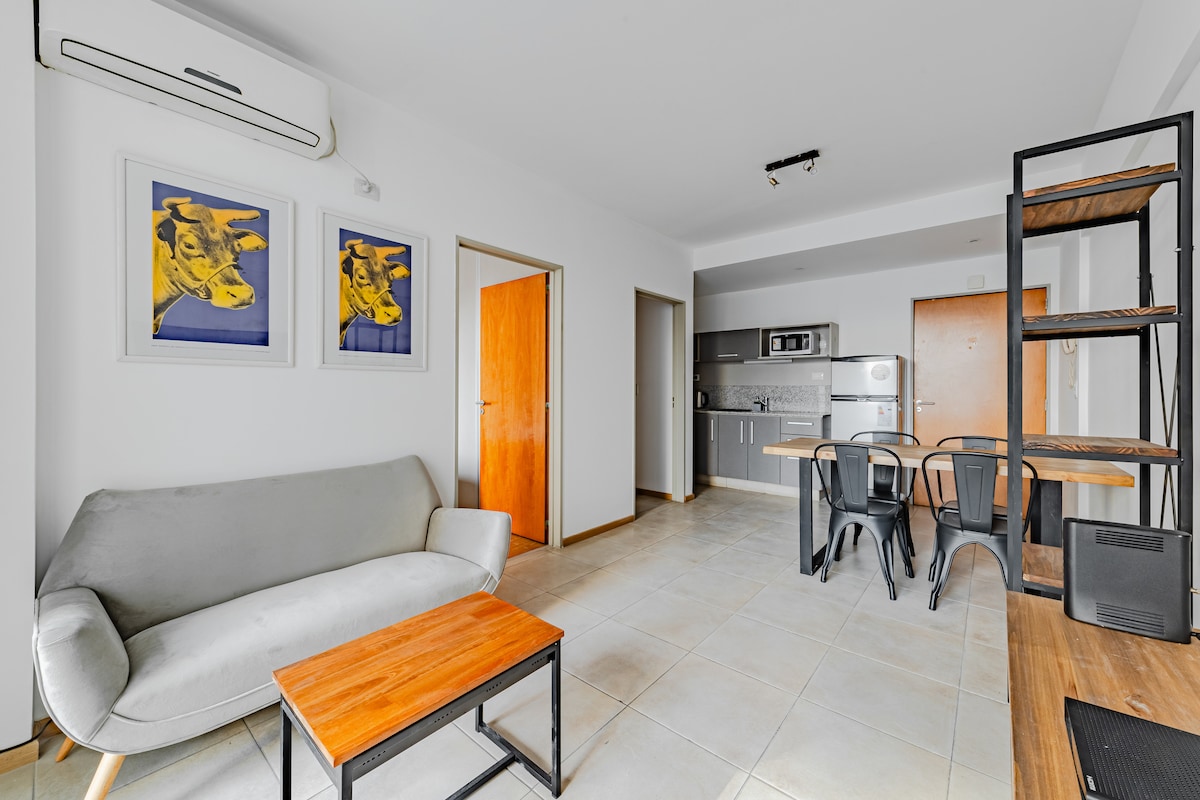The living area features a comfortable sofa alongside a wooden coffee table. Framed artwork adorns the wall, while a dining space with a table and black chairs is visible. An open kitchen is equipped with modern appliances, including a microwave and stove.