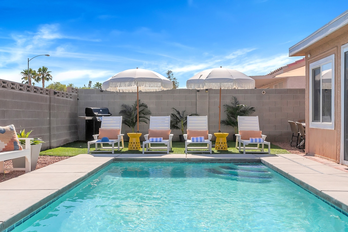 A private pool offers a refreshing retreat, surrounded by lounge chairs and vibrant umbrellas. A grill stands nearby, complemented by grass and plants for added greenery. The space is designed for relaxation and enjoyment under a clear blue sky.