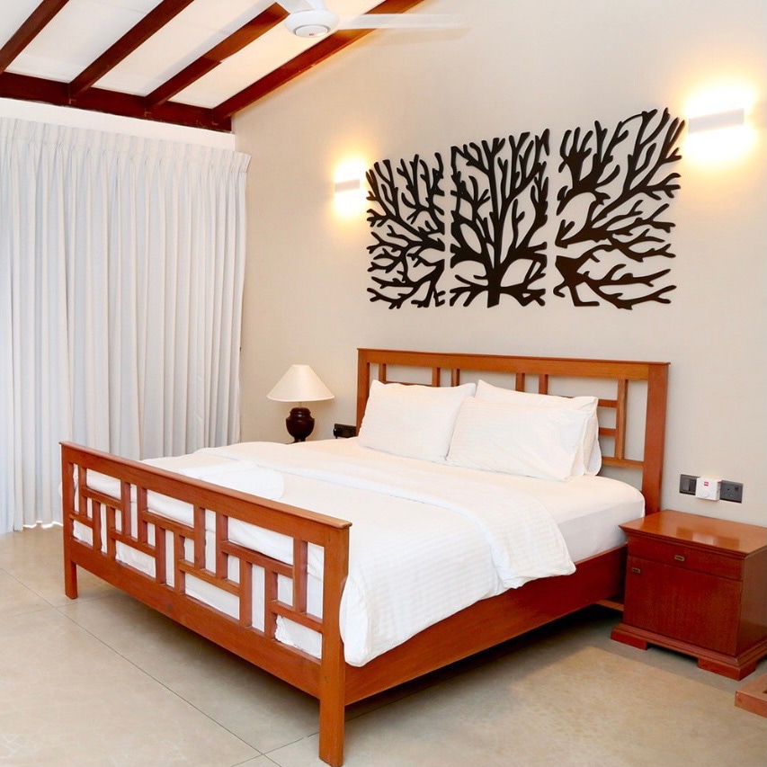 The sleeping area features a wooden bed with a clean white bedding set. A decorative wall art composed of coral shapes is displayed above the bed. A nightstand with a lamp is placed next to the bed, and soft curtains partially cover a window.