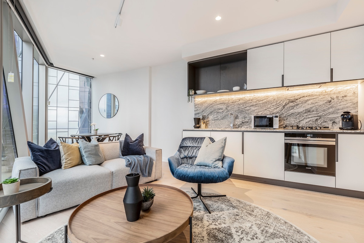 An open-plan lounge area features a comfortable grey sofa adorned with cushions in various tones. A round dining table sits nearby. The sleek kitchen showcases modern appliances and a marble backsplash, while natural light flows through large floor-to-ceiling windows.