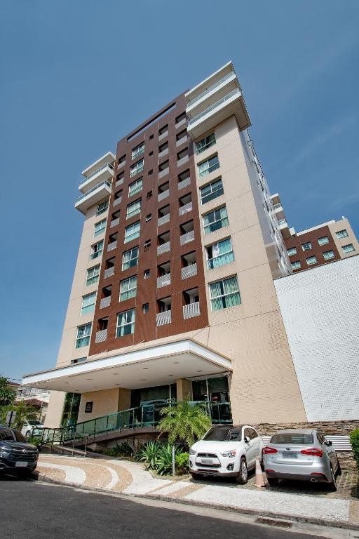 The façade of a modern multi-story building is displayed, showcasing a combination of light-colored and dark wooden accents. The entrance features a covered area with a sloped pathway leading up to it, surrounded by green plants and parked vehicles.