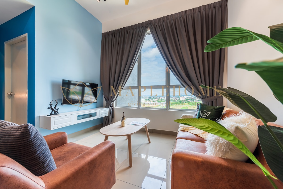 A cozy living area is presented with two brown leather sofas positioned around a light wooden coffee table. A flat-screen television is mounted on the wall opposite the seating, and large windows with dark curtains allow natural light to brighten the space.