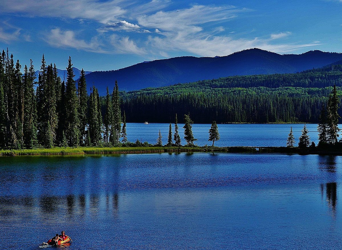 A calm lake reflects surrounding greenery and distant mountains under a clear blue sky. A small boat glides across the water, creating gentle ripples. Pine trees are seen along the shore, contributing to a natural, serene landscape.