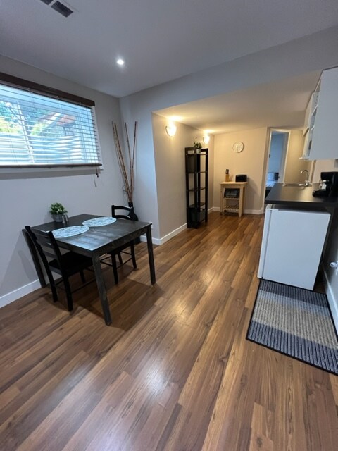 A cozy dining area features a small wooden table set for two with simple tableware. A black shelf stands nearby, and light streams in through a window with blinds, illuminating the warm, wooden floors. The kitchenette area, equipped with essential appliances, is visible in the background.
