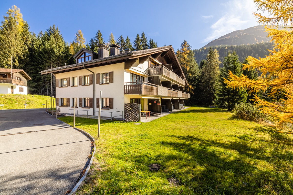 A multi-story building surrounded by trees and a grassy area is featured. The structure includes multiple balconies with wooden railings. A gravel path leads to the entrance, and the backdrop presents a scenic view of the mountains and clear blue sky.