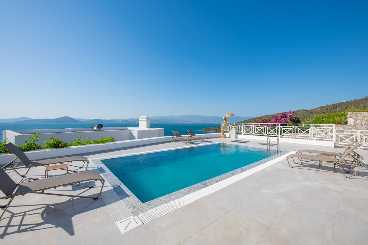 A swimming pool is surrounded by several lounge chairs, with a scenic view of the distant mountains and sea. Bright blue skies create a serene atmosphere, while vibrant greenery decorates the nearby terrace.