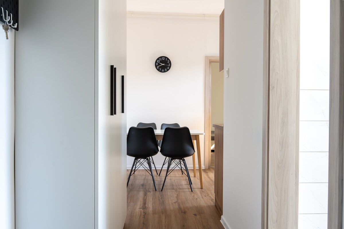 A brightly lit hallway leads to a dining area featuring a wooden table surrounded by four black chairs. A simple wall clock hangs above, and light wooden flooring extends throughout the space, creating a sense of openness.