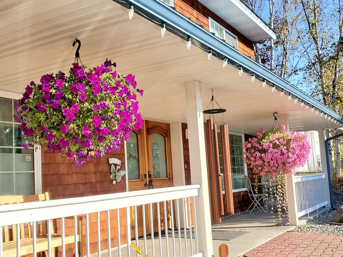 A welcoming porch is adorned with vibrant hanging flower baskets in shades of pink and purple. Comfortable seating options are available, along with a small table for outdoor relaxation. The entrance is framed by natural wood accents and soft lighting, creating an inviting ambiance.