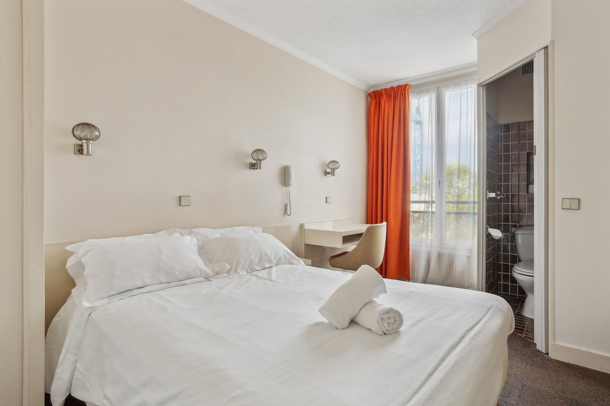 A well-lit room features a spacious bed dressed in fresh linens, complemented by neatly arranged towels. Natural light floods the space through a window adorned with sheer curtains. A small desk and a wall-mounted lamp are situated nearby, enhancing functionality.
