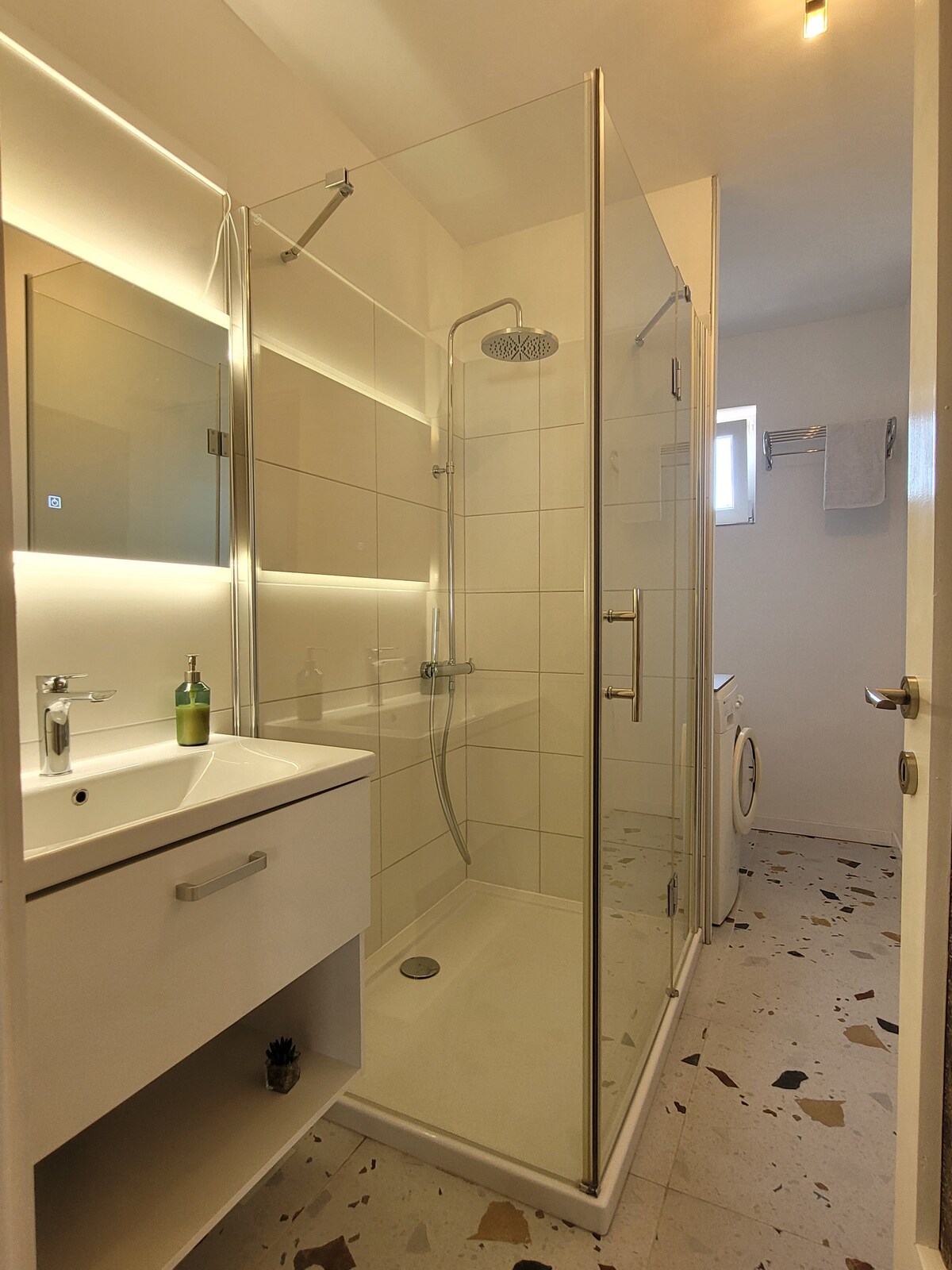 A modern bathroom is presented with a glass-enclosed shower featuring a rainfall showerhead. A sleek vanity with a mirror and an additional towel bar is visible. Natural light enters through a small window, while a washing machine is discreetly placed in the corner.