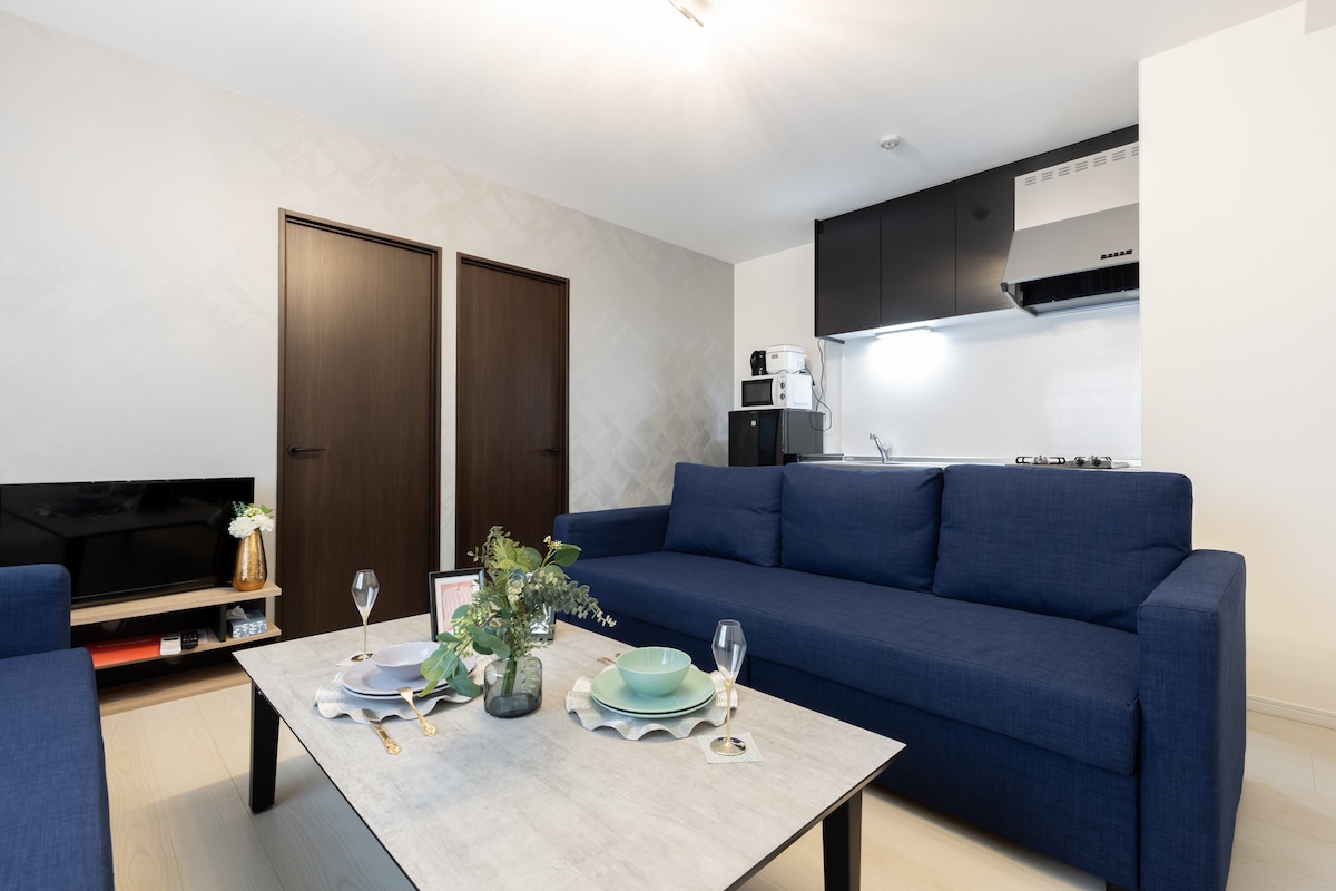 A spacious living area features a large blue sectional sofa surrounding a light-colored coffee table, set with tableware and a floral centerpiece. Two wooden doors are visible, leading to private areas, while the kitchen area, equipped with modern appliances, is seen in the background.