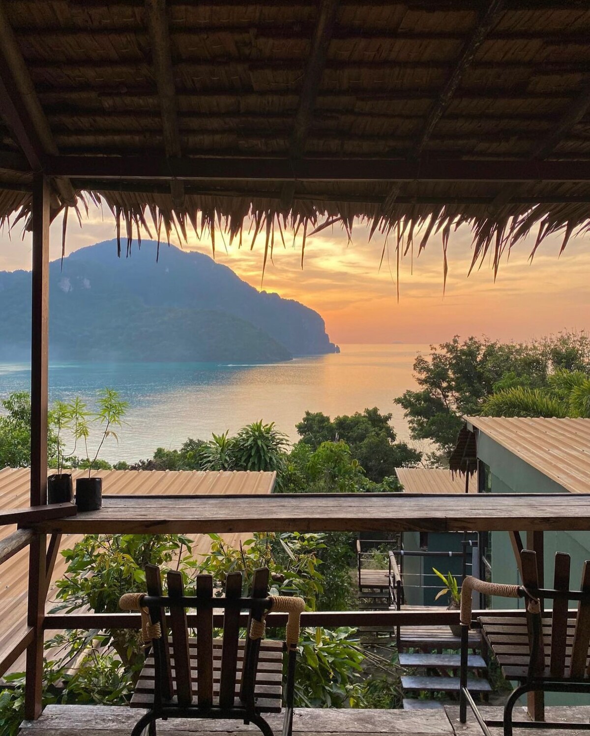 A covered outdoor terrace offers two wooden chairs facing a serene ocean view, framed by lush greenery. The backdrop features a mountainous silhouette under a soft, colorful sky, suggesting a tranquil sunset over the water.