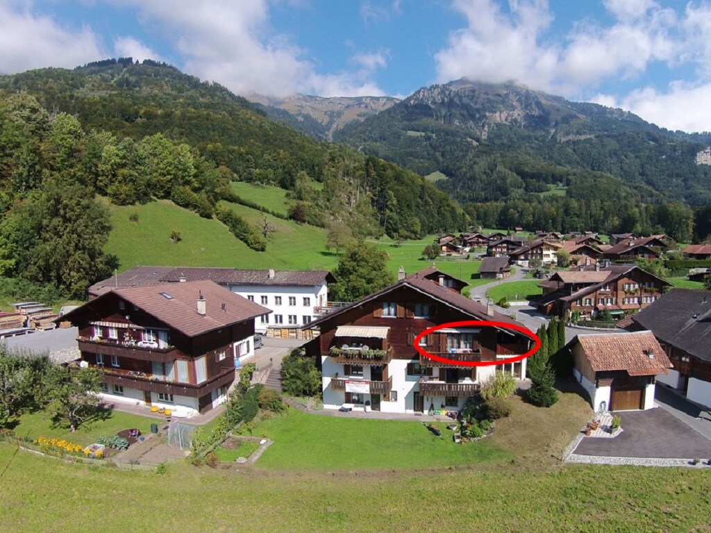An aerial view of a charming village surrounded by lush green hills. The balcony of the highlighted apartment is visible, overlooking the tranquil landscape. Nearby buildings showcase traditional architecture, with gardens and open spaces creating a serene environment.