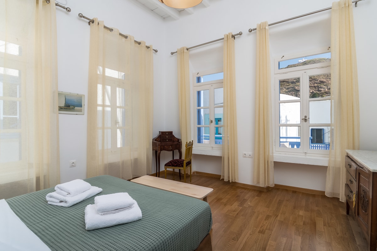 A bright sleeping area is featured, with large windows allowing natural light to fill the space. Light, sheer curtains frame the windows. A spacious bed is complemented by neatly arranged towels. A small wooden table and a chair add to the minimalist decor.