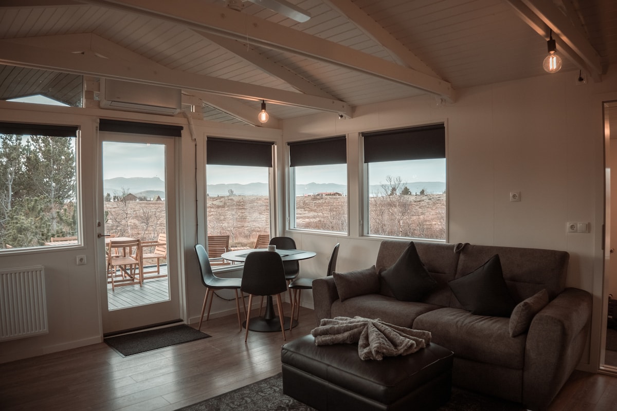 The living area features a comfortable sofa paired with dark cushions. Large windows offer expansive views of the surrounding landscape. A dining table with four chairs is positioned nearby, complementing the bright, airy space illuminated by hanging light fixtures.