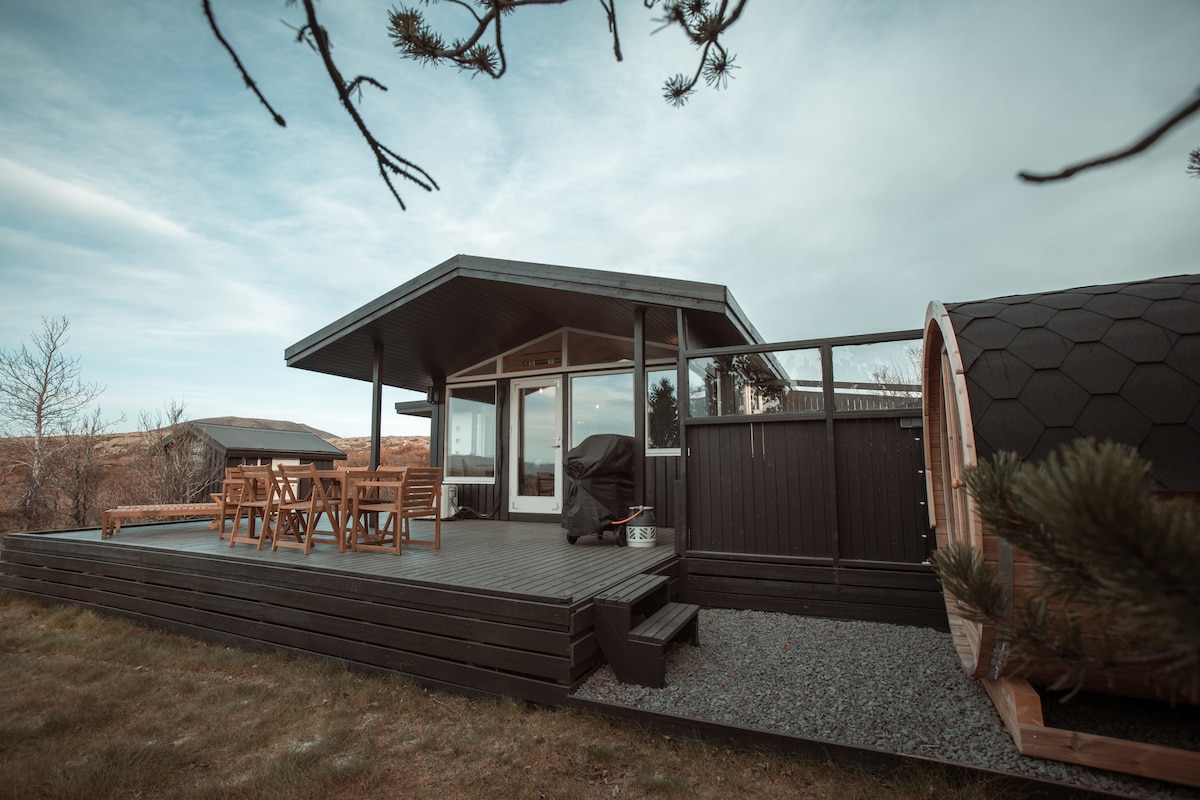 A sleek cabin exterior features a spacious wooden deck furnished with a dining table and chairs, situated beside a wood-fired sauna. Large windows enhance natural light, while views of the surrounding landscape are apparent. A gravel pathway leads from the deck to the yard.