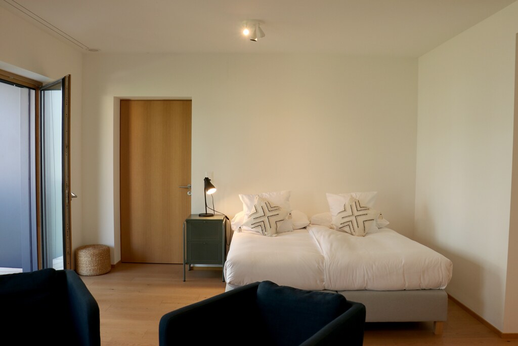 A bright sleeping area features a spacious bed with decorative pillows. A bedside table with a lamp provides soft lighting. A door leads to the balcony, while a basket and two chairs in a cozy nook enhance the room's functionality.