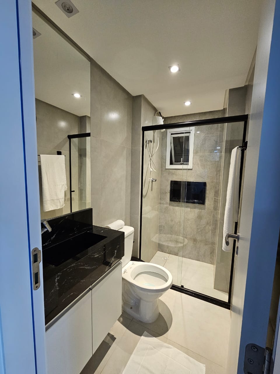 A modern bathroom features a sleek, dark stone countertop with a single basin sink. A glass-enclosed shower is visible, showcasing tiled walls and a showerhead. White towels hang on a rack, while a window provides natural light. The space is well-lit with recessed lighting.