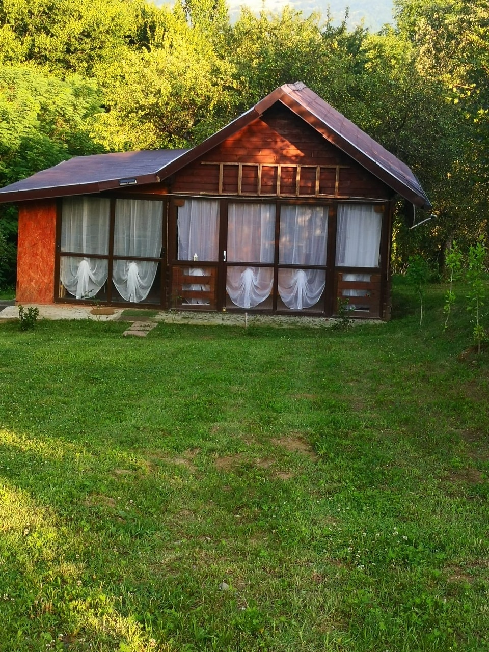 A wooden cabin is situated in a lush green area, featuring large windows with sheer curtains. The sloped roof is complemented by a natural setting, with grass and trees surrounding the structure. A stone pathway leads to the entrance, enhancing the tranquil ambiance.